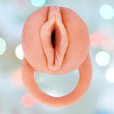 A white silicone product, The Great Extender 2-in-1 Penis Extender & Masturbator from Nasstoys, featuring an elegant design with a circular handle, is artfully showcased against a softly blurred background with pastel-colored bokeh lights.